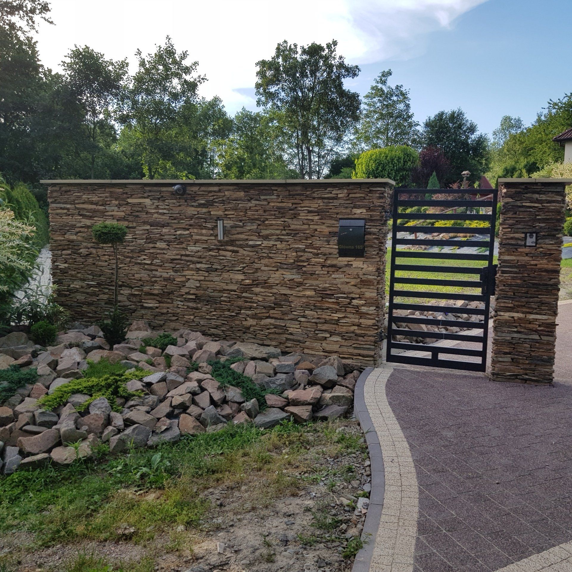 Stone wall with black metal gate and mailbox, surrounded by landscaping, under a blue sky.