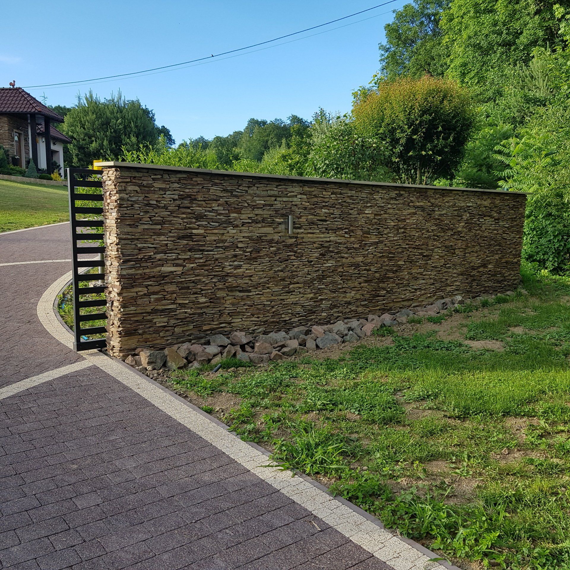 Stone wall with black metal gate next to a paved driveway and green lawn.