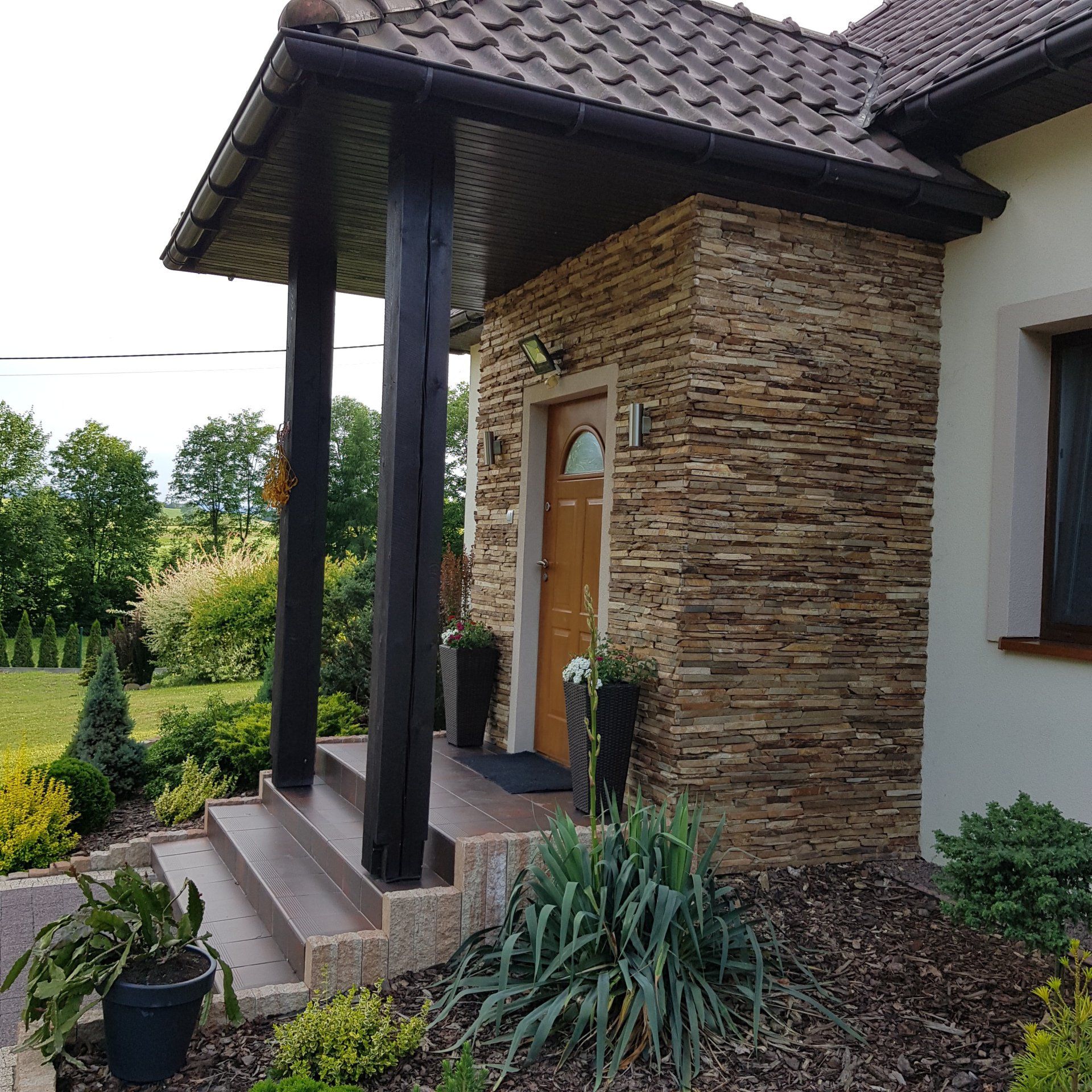 House entryway with stone facade, wooden posts, and brown door; steps lead up to the entrance.