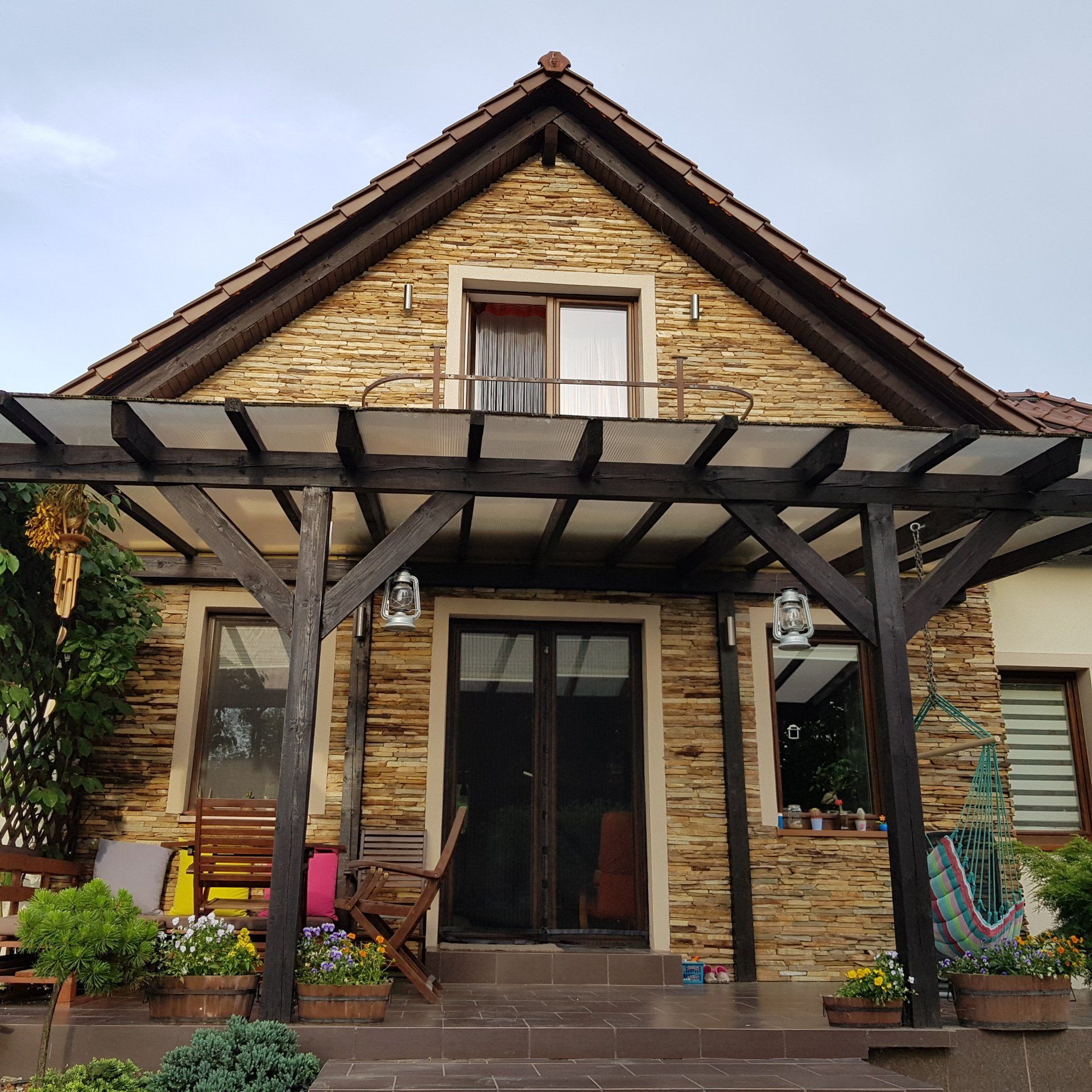 Stone house with pergola-covered porch, flower pots, and wooden furniture.