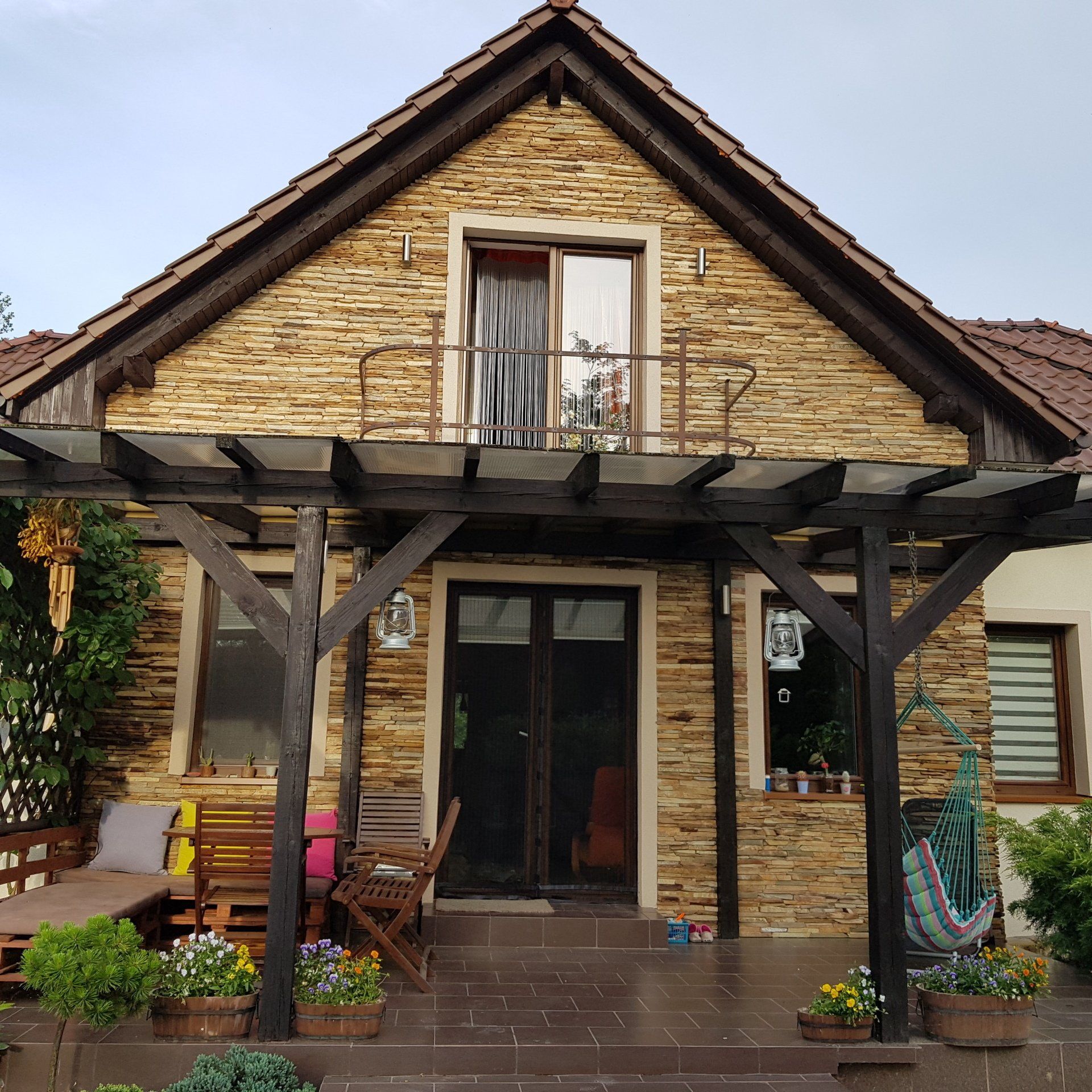 Stone-walled house with wooden pergola, balcony, and front steps. Plants, outdoor furniture, and hammock decorate the porch.