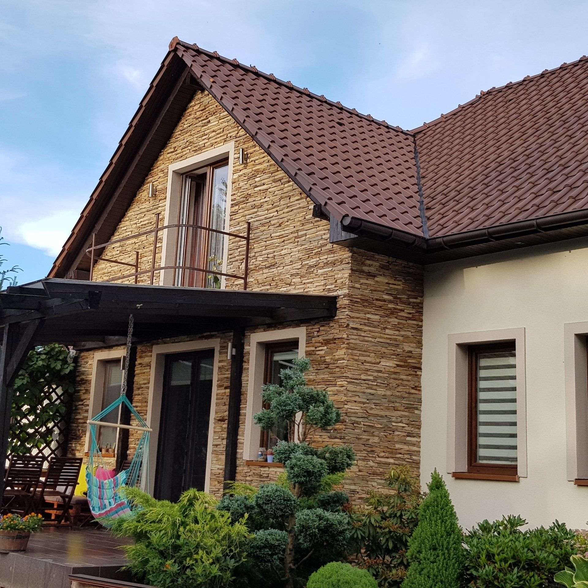 Stone-clad house with brown roof and balcony, flanked by bushes and pergola with swing on a sunny day.