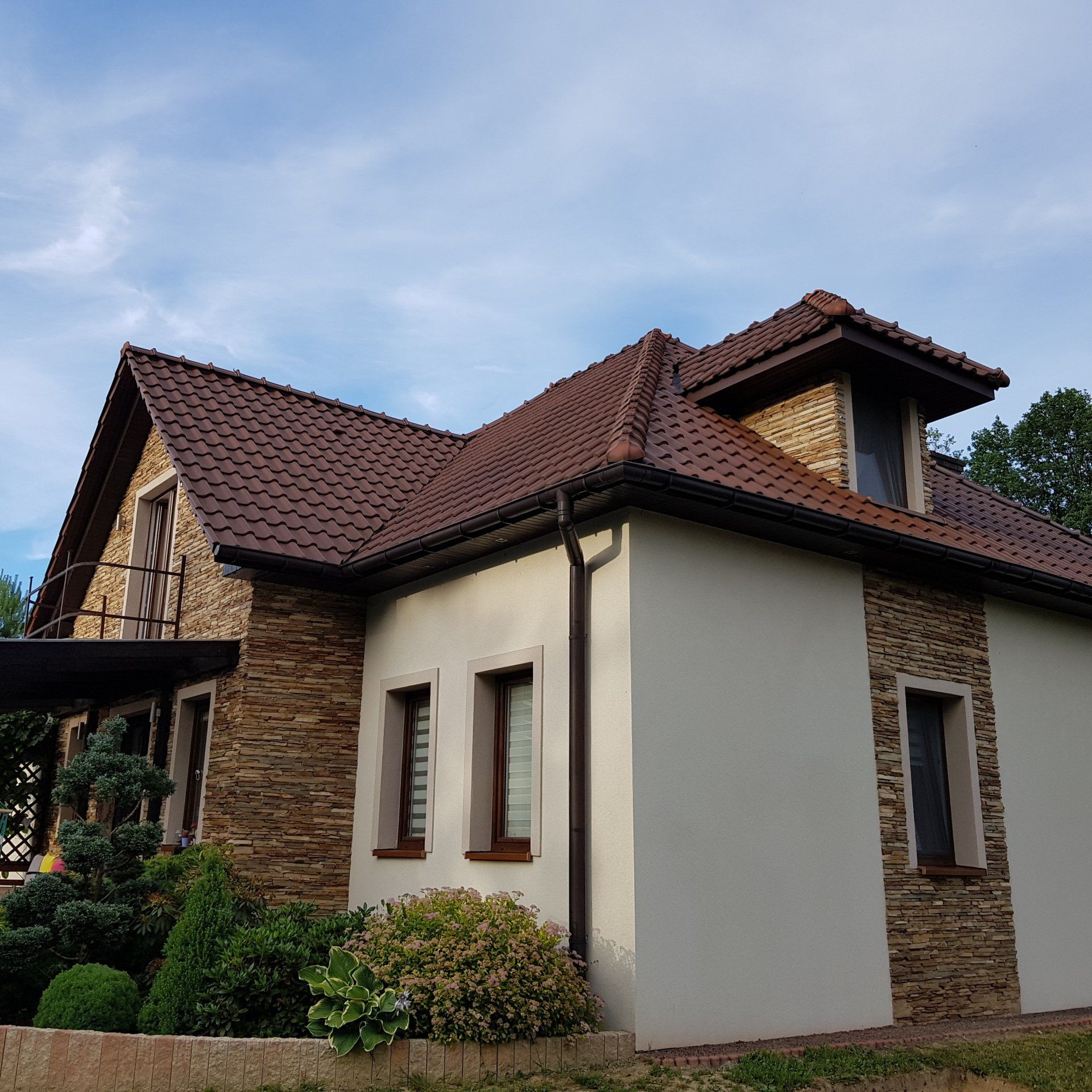 House with brown roof, stone accents, and beige siding against a cloudy blue sky. Landscaping in front.