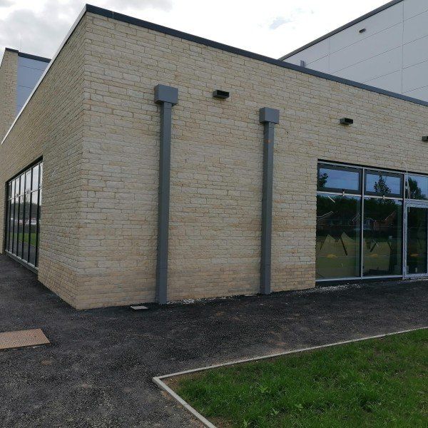 Beige brick building with glass windows, gray pipes, and a paved walkway next to grass.