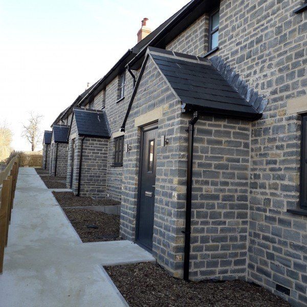 Row of stone houses with dark doors, under black roof overhangs, concrete path in front.