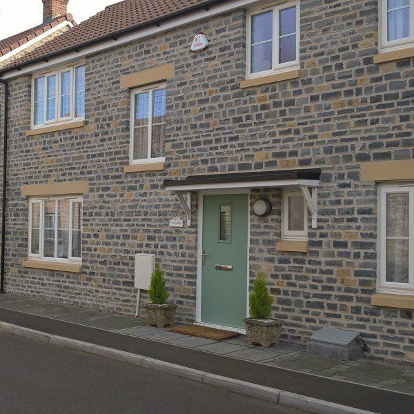 Stone house with a green door and small awning. White-framed windows, two small potted trees by the door.