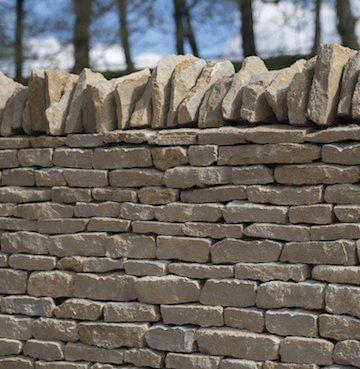 Stone wall with uneven top, built with light-colored, rectangular stones, set against a blurred background of trees and sky.