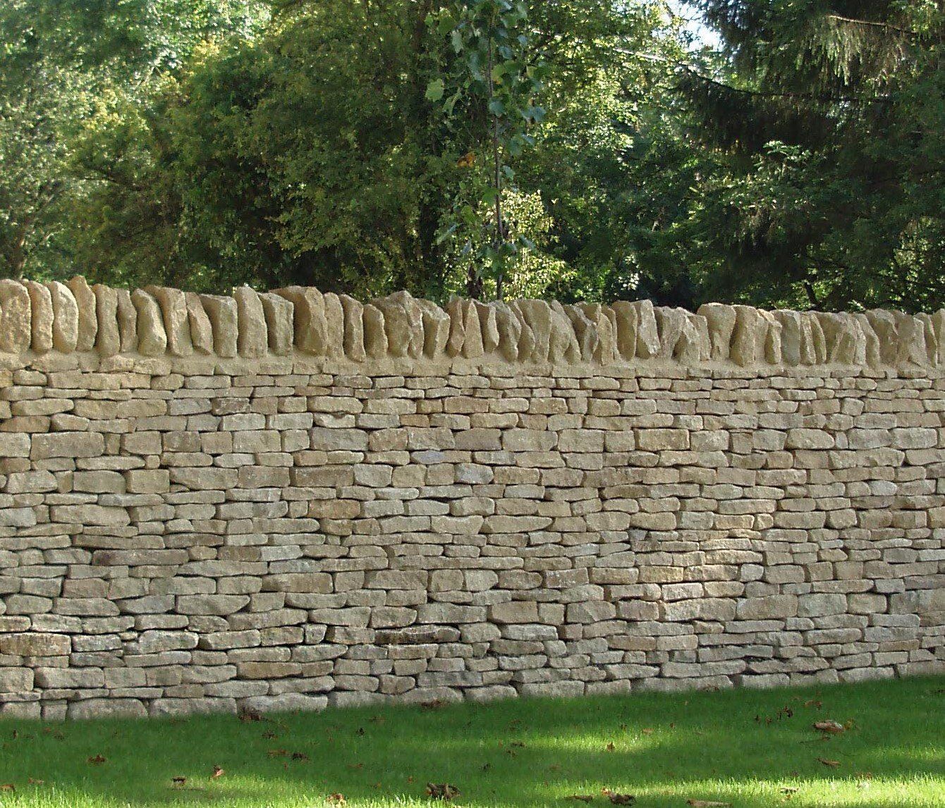 Stone wall with rough-edged top, built from rectangular blocks. Green grass in foreground, trees behind.