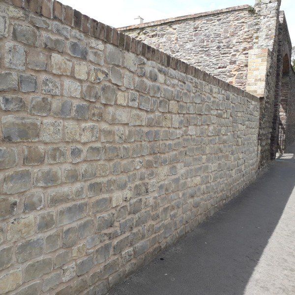 Stone wall next to a paved path, with a brick top and an arched structure visible.