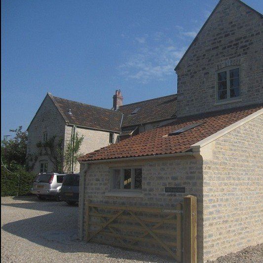 Stone building with red-tiled roof, wooden gate, and gravel driveway under a blue sky.