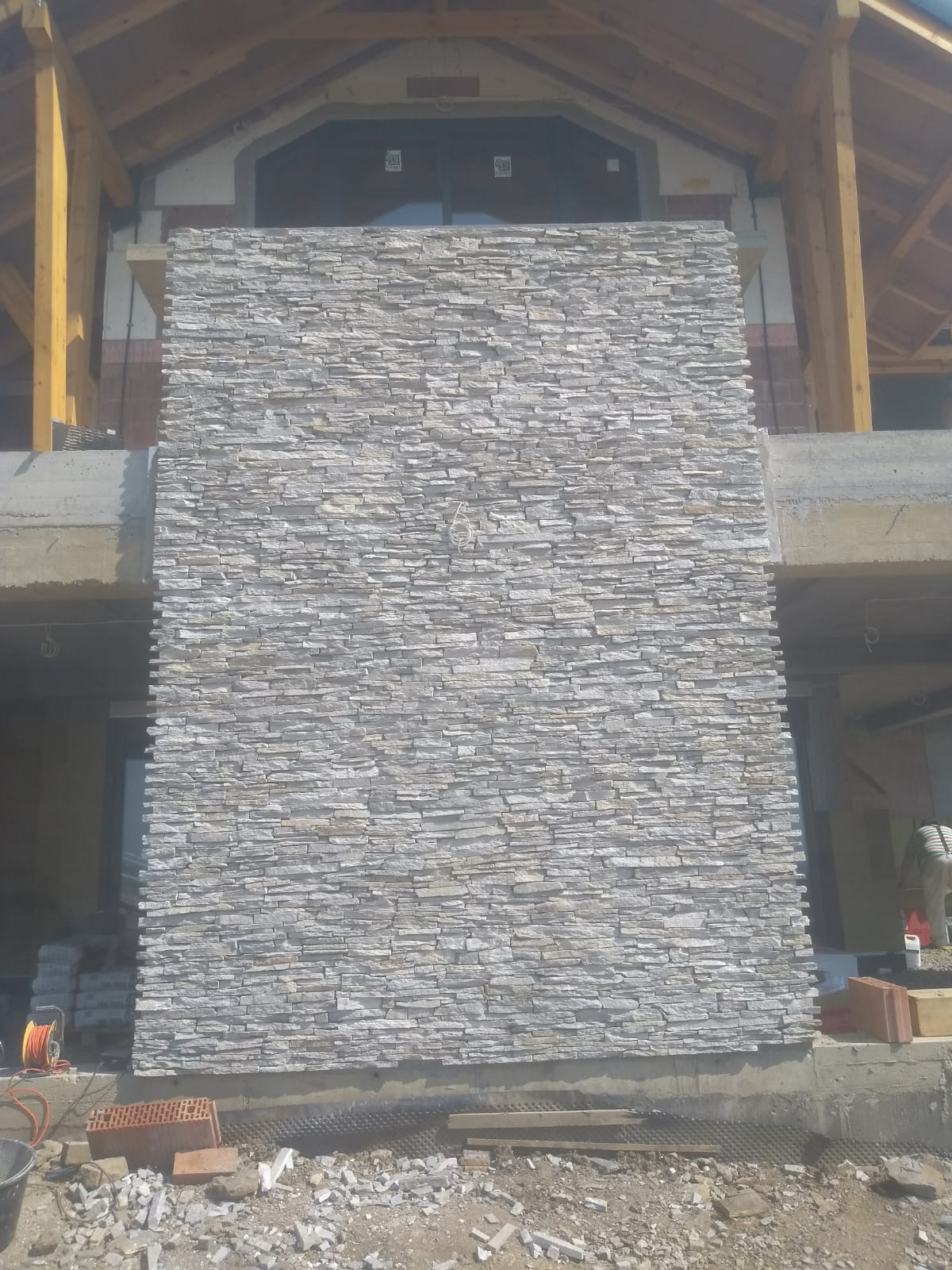 Stone veneer wall under construction; building with wooden frame and concrete base in background.