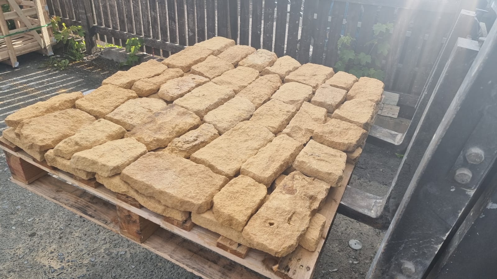 Pallet of light brown bricks outdoors in front of a wooden fence, with sunlight visible.