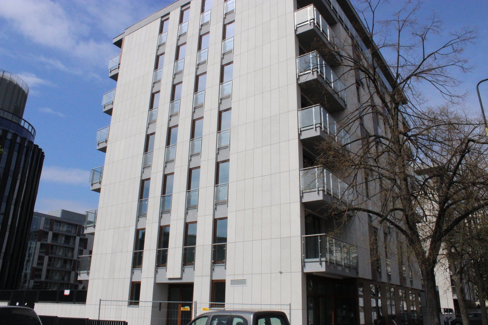 Modern apartment building, light gray facade, balconies, blue sky in the background, trees on the right.