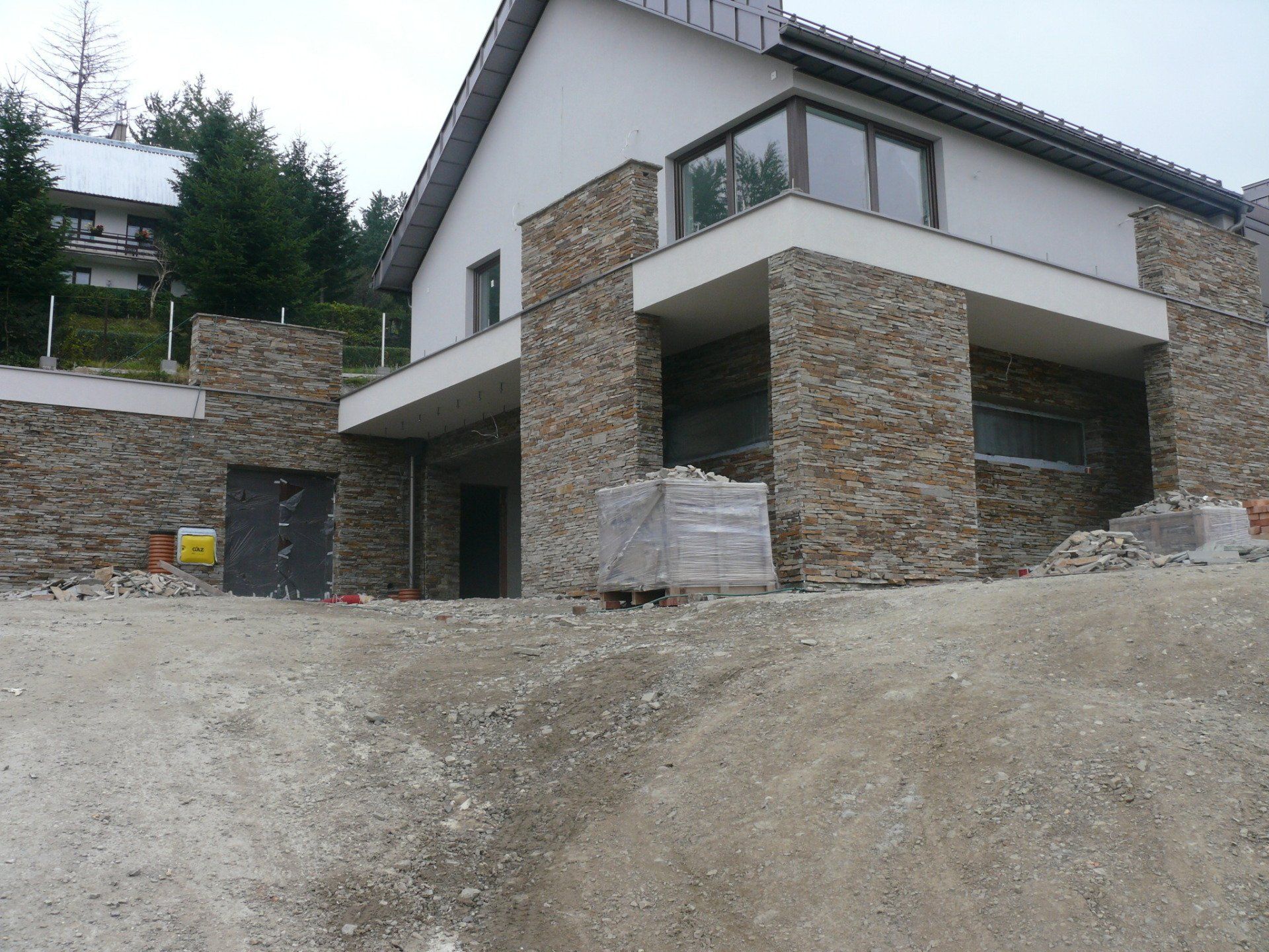 Modern house under construction with stone facade and gravel yard.