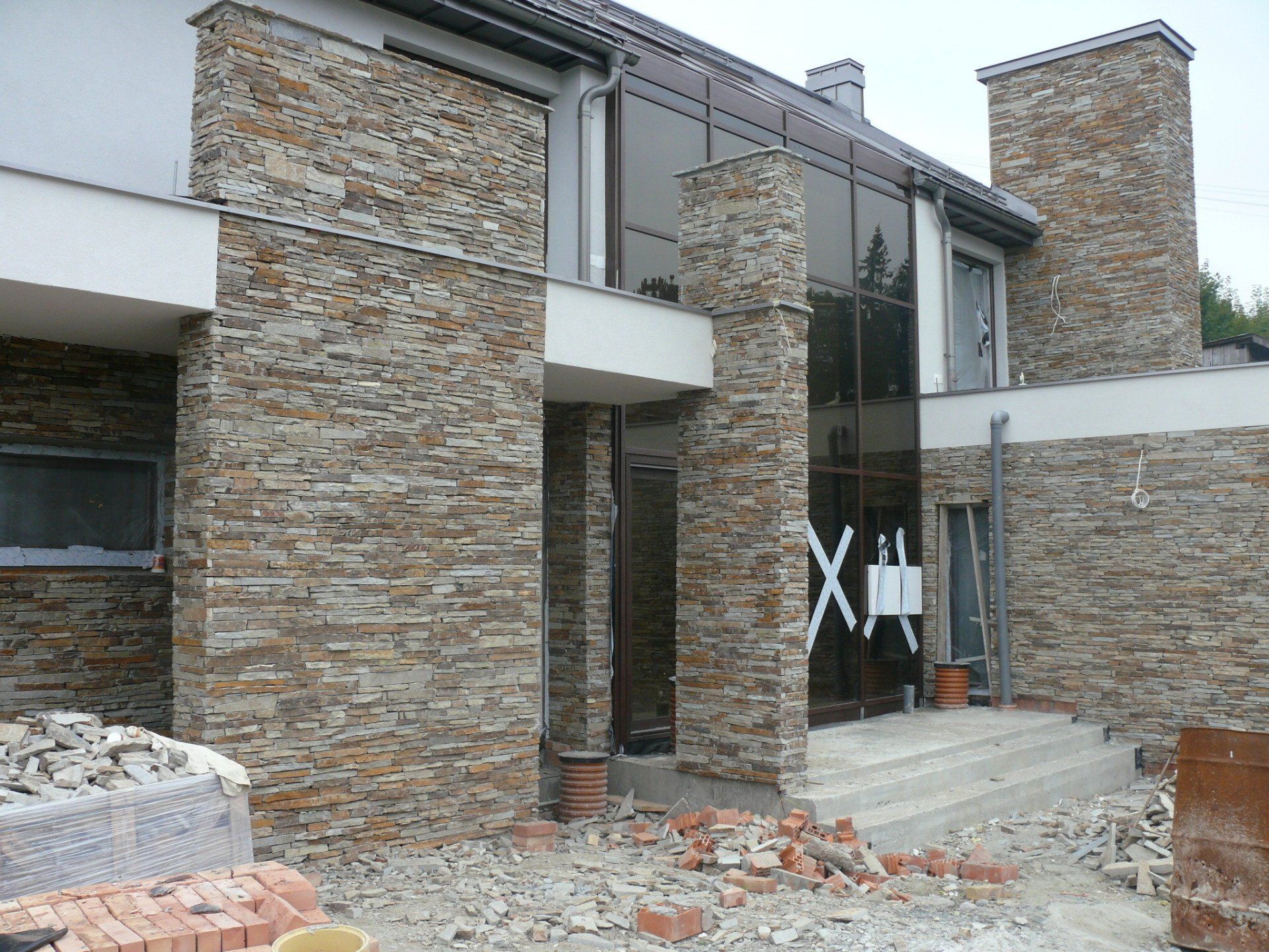 Modern house exterior under construction, stone facade, glass entrance, brick debris.