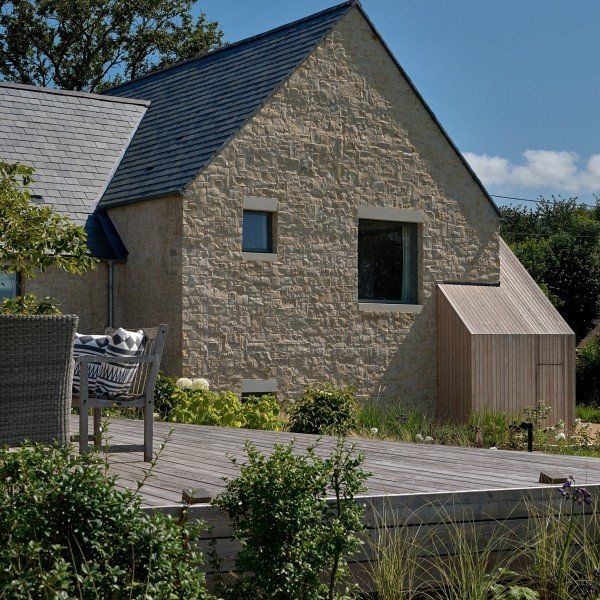 Stone house with wooden deck, dark roof, small windows, and a wooden side extension against a blue sky.