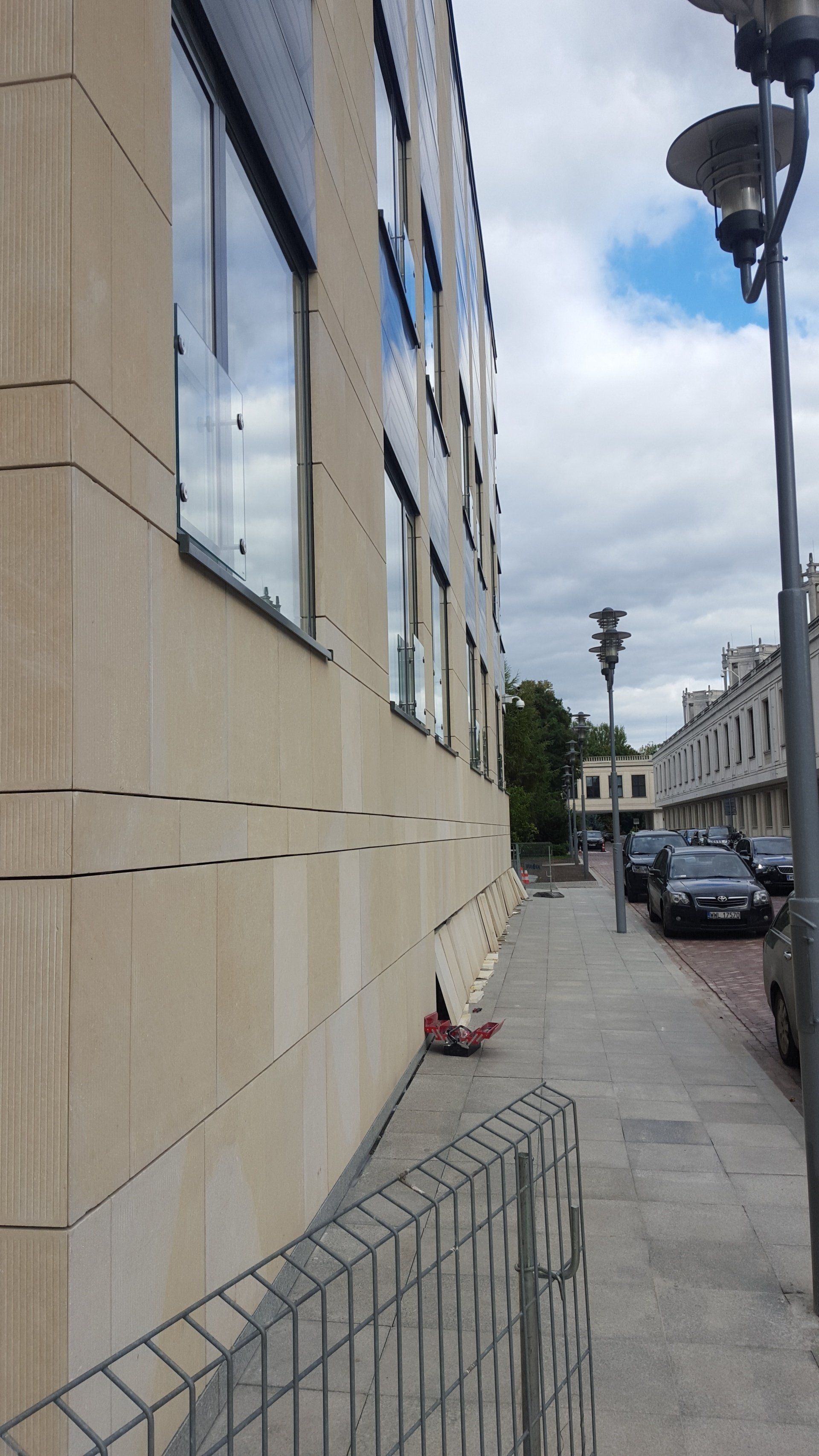Building exterior with windows, sidewalk, street, cars, and a wire fence on a cloudy day.
