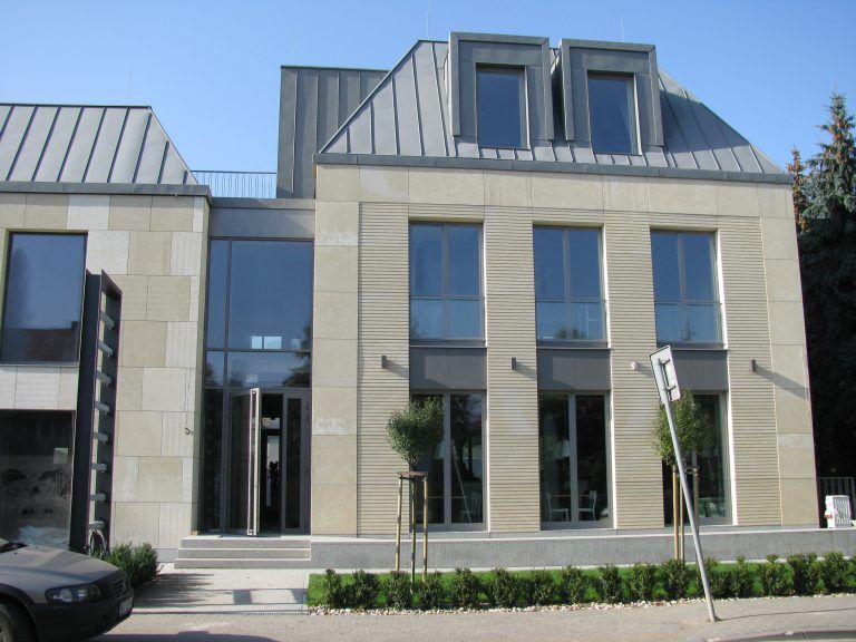 Modern two-story building with light stone facade, glass doors, and gray metal roof under a blue sky.