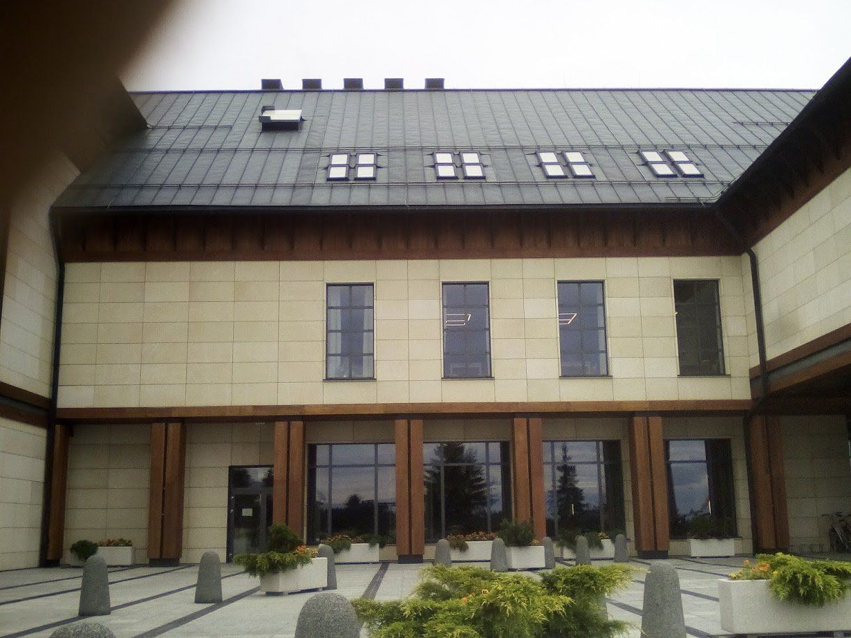 Stone building with dark roof, windows, and light-colored pillars. Entryway with plants in planters.