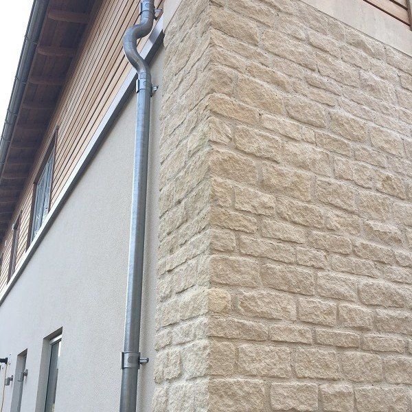 Corner of a building with stone facade, light-colored walls, and a metal downspout.