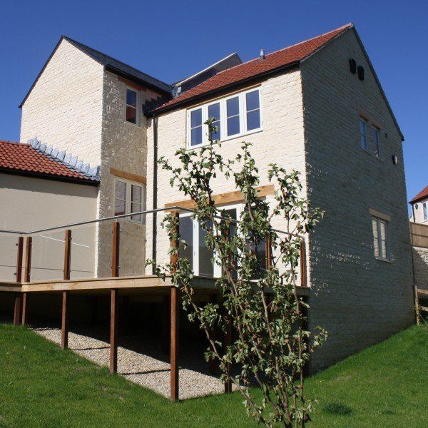 Two-story brick house with a wooden deck and red-tiled roof, set on a grassy hill under a blue sky.