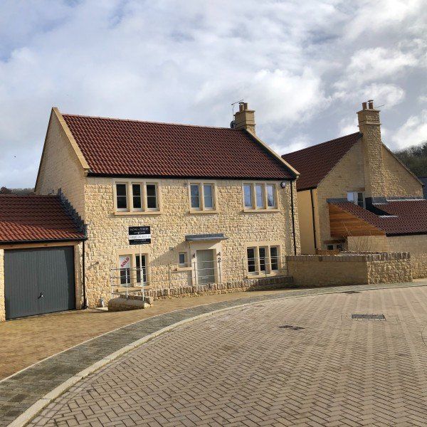Two-story beige stone houses with red roofs sit on a paved road, under a cloudy sky.