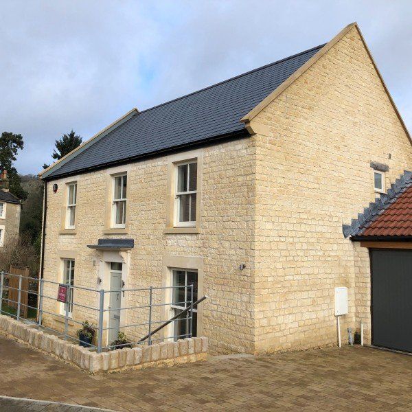 Two-story stone house with dark roof and white window frames, near a paved driveway.