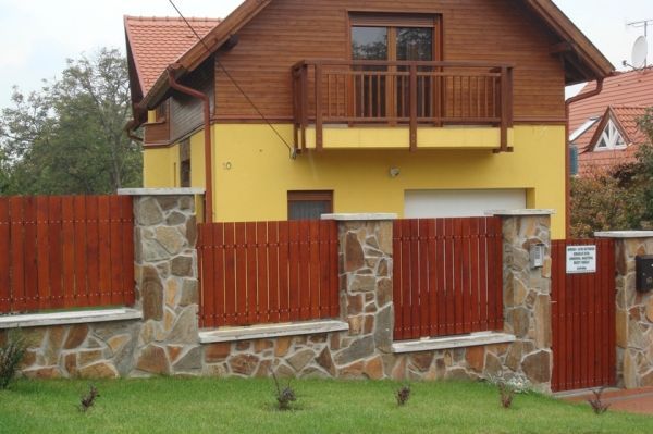 Yellow house with a brown wooden balcony, red tiled roof, and a wooden fence with stone columns.