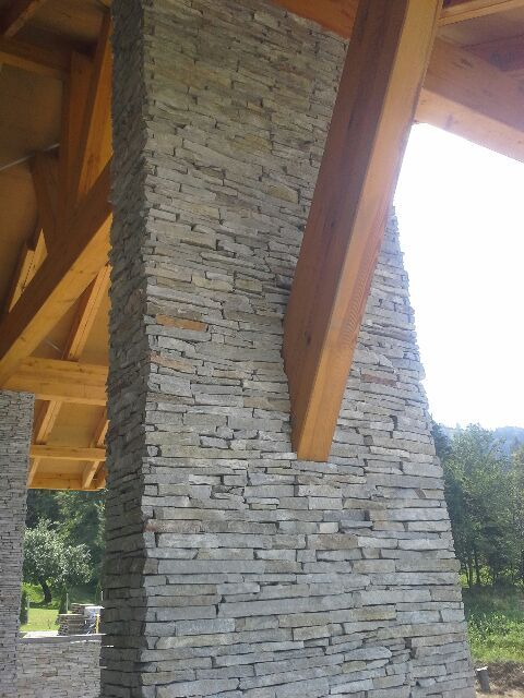 Stone-clad pillar supporting a wooden beam structure; exterior view with trees in the background.