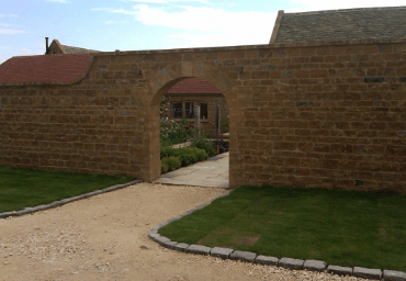 Stone archway entrance with tan gravel path and green lawn. Building with red roof visible.