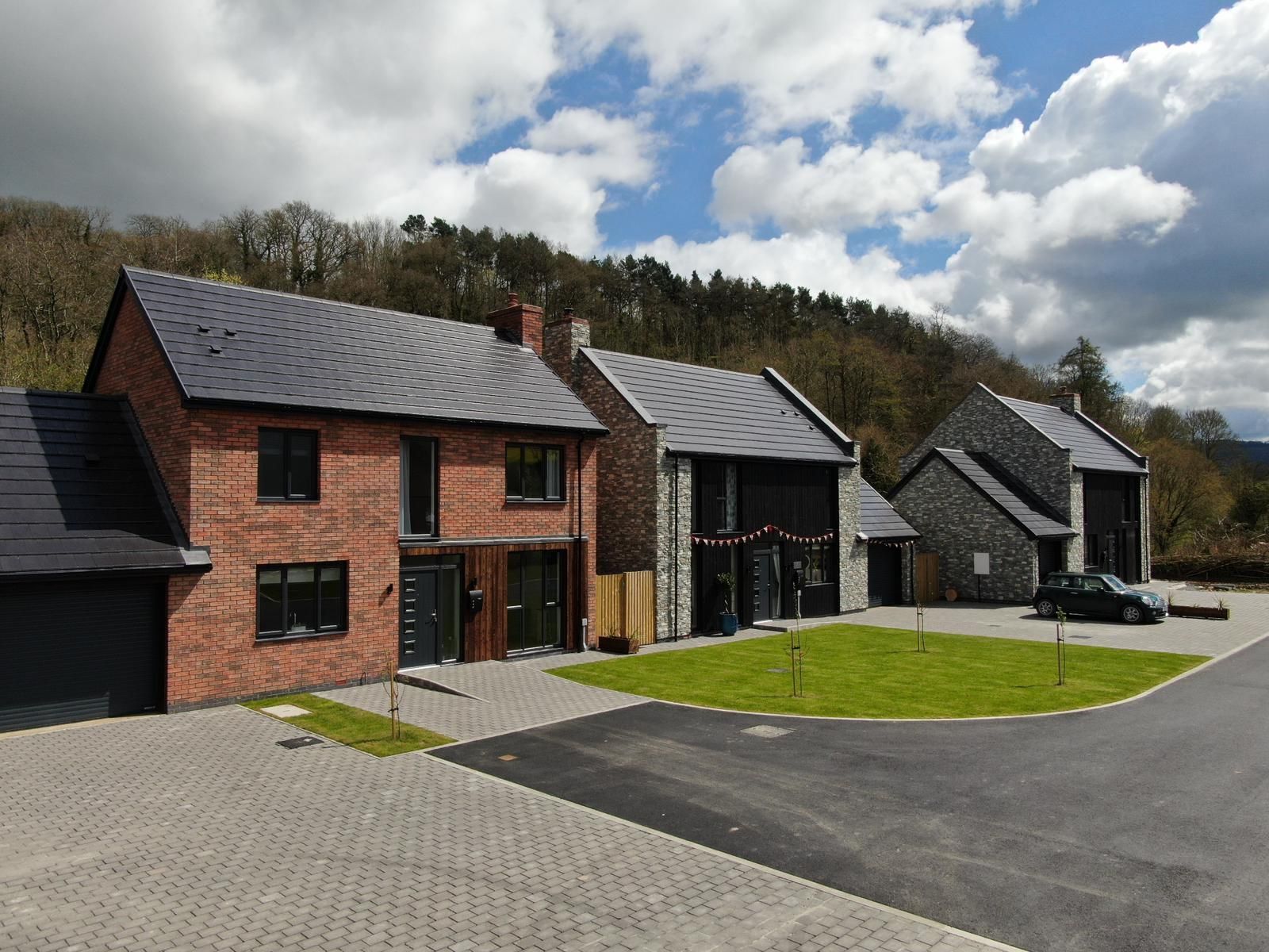 Row of modern houses with brick, slate, and stone exteriors, set against a hillside under a cloudy sky.