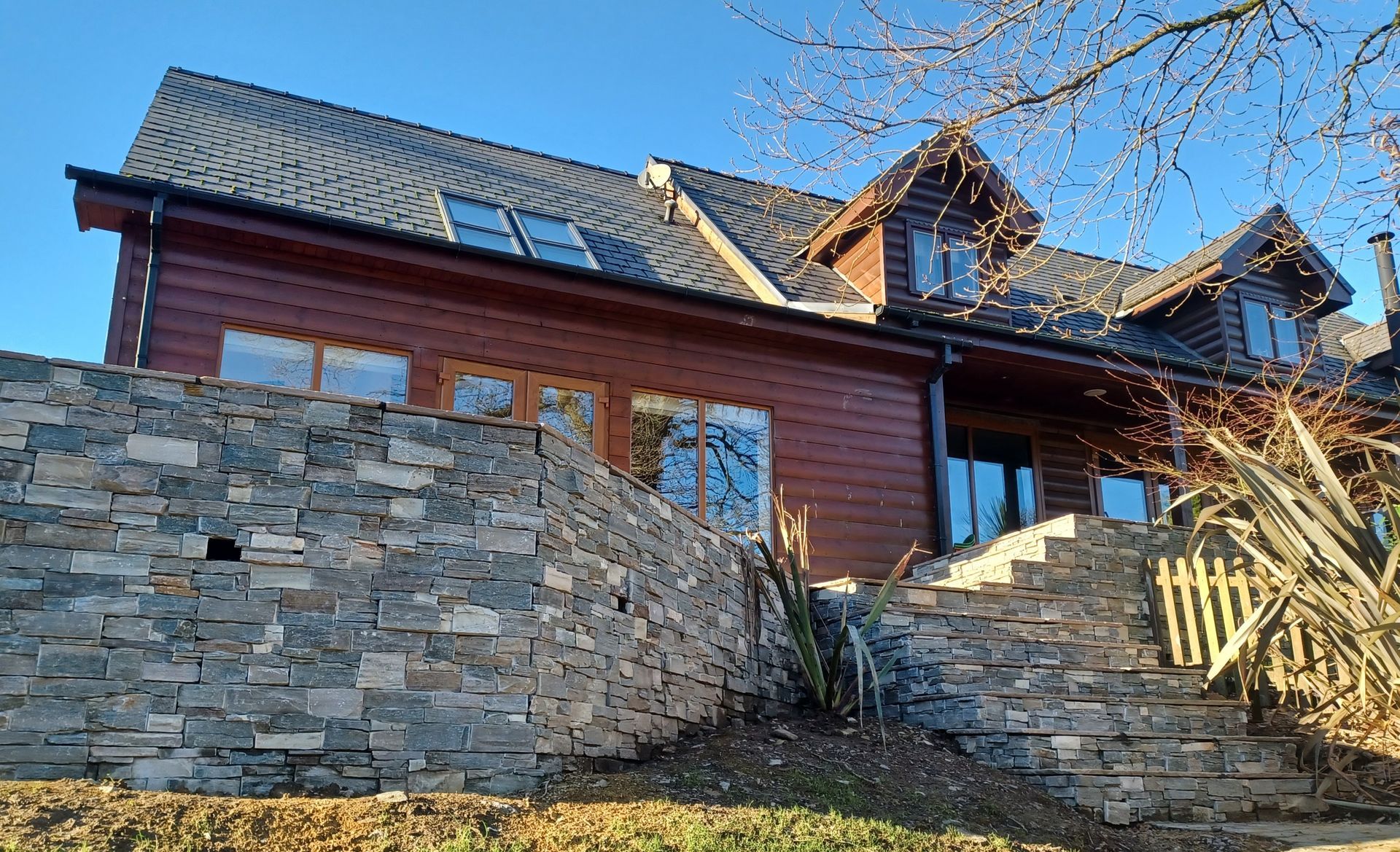 Brown wooden house with stone wall and dark roof against a blue sky.