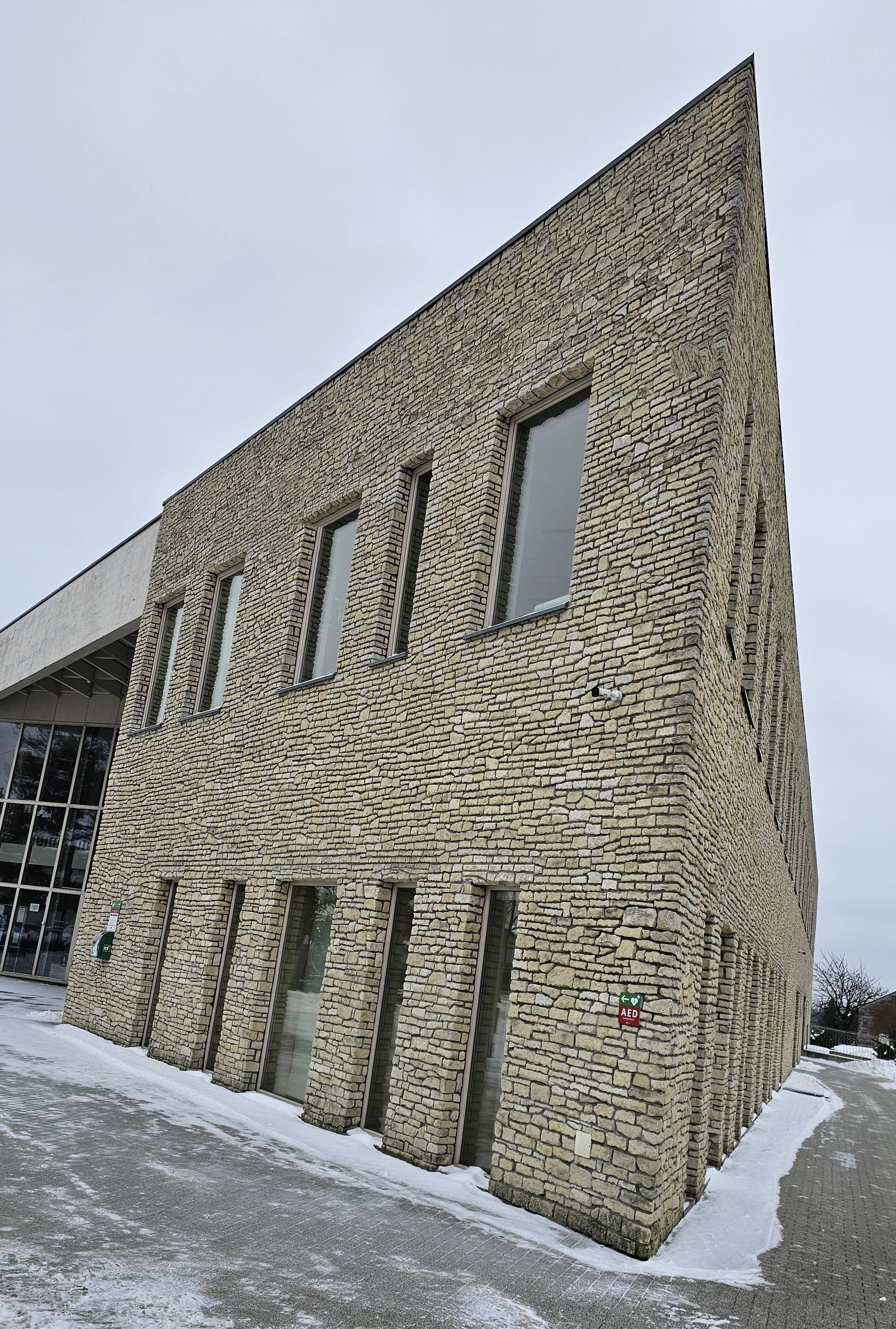 Angular building with textured brick facade and windows, set against a cloudy sky with patches of snow.