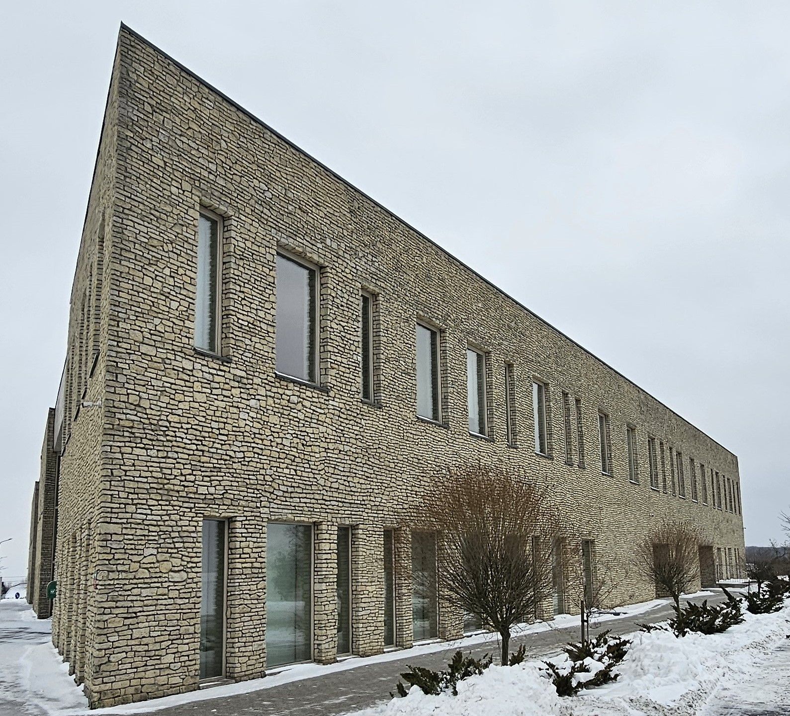 Triangular stone building with multiple windows, surrounded by snow and sparse bushes.