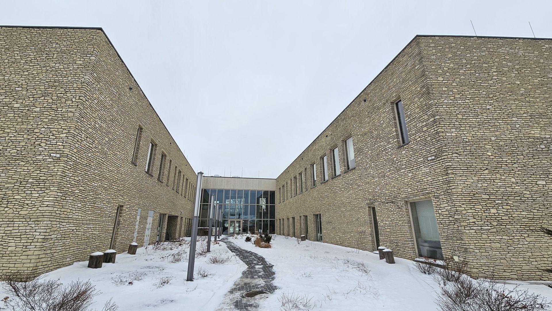 Two-story beige brick buildings form a V shape, leading to a glass entrance, snowy ground.