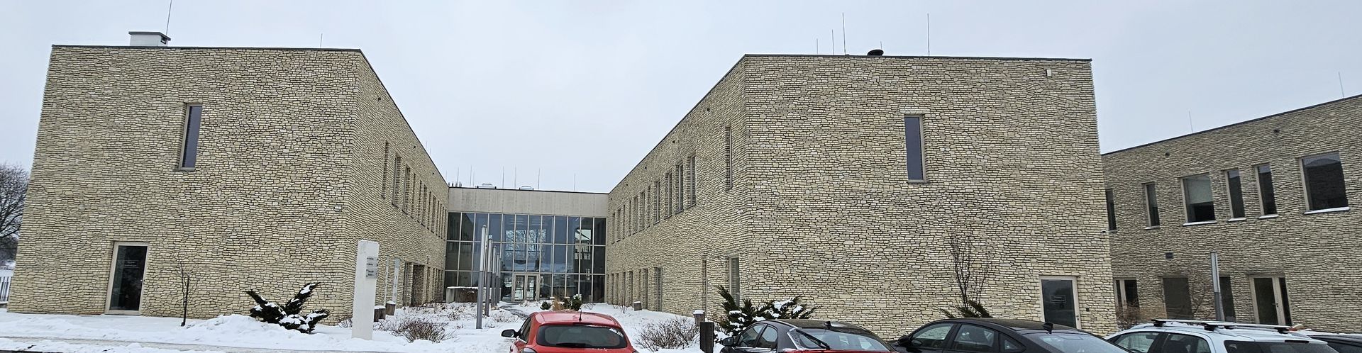 Stone building with a glass entrance, snowy ground, and parked cars. Overcast sky.