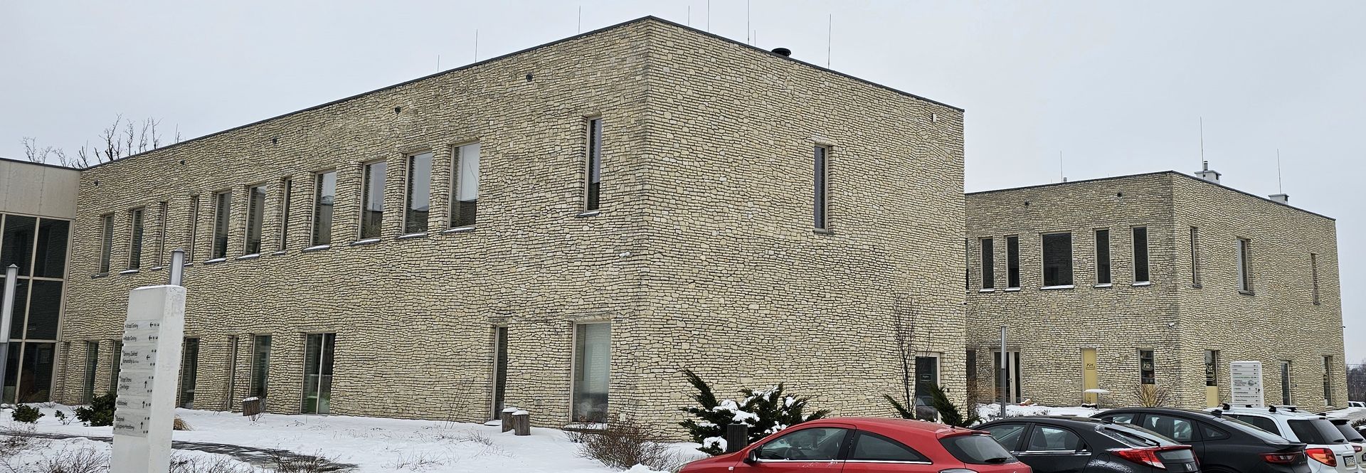 A large building with textured stone facade on a snowy day; parked cars in the foreground.