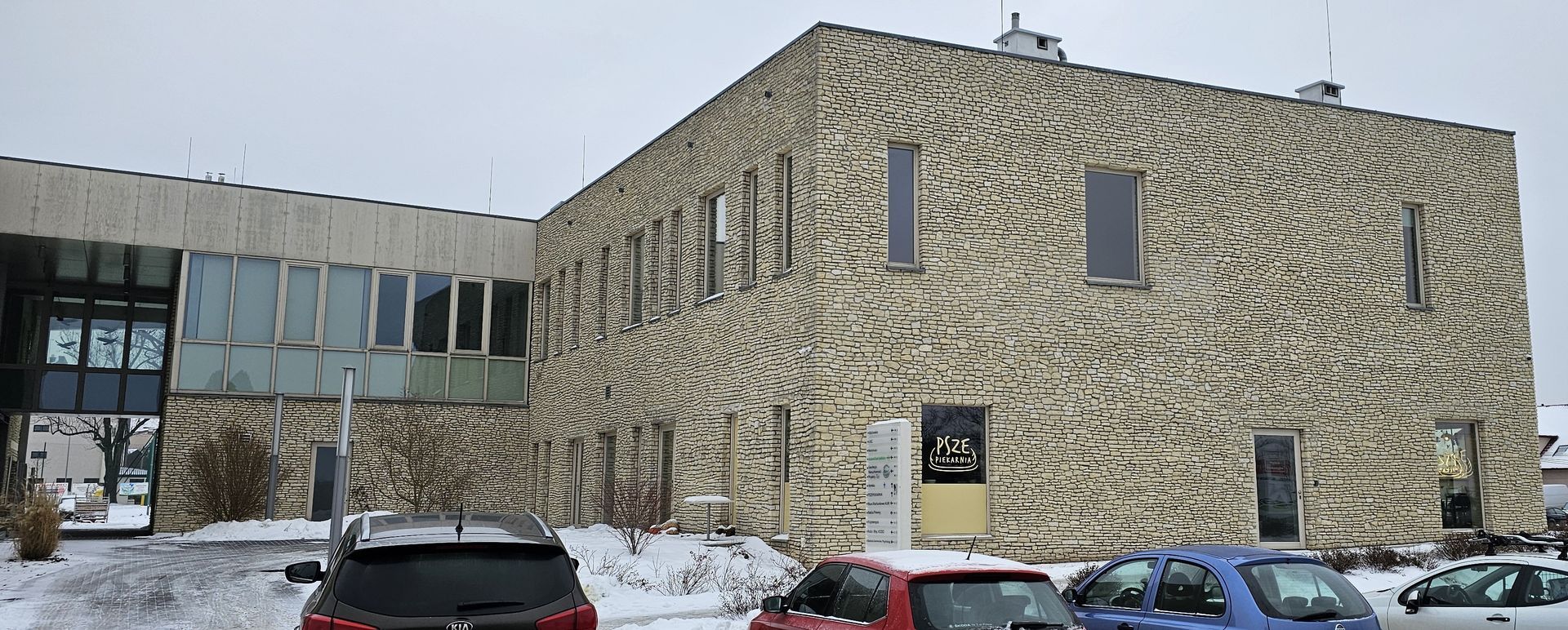 A two-story building with rough stone facade, snow on the ground, and cars parked in front.