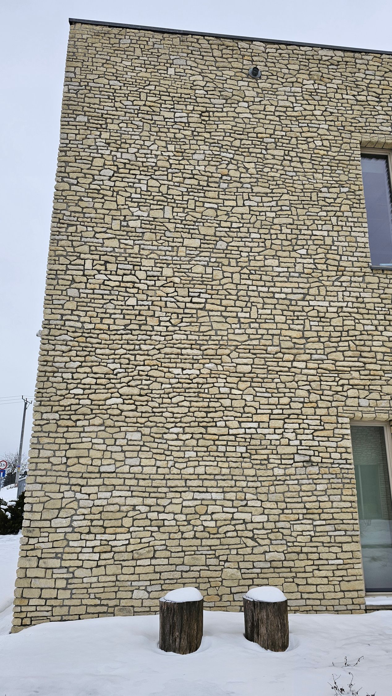 Stone-clad building exterior with stacked horizontal stone pattern, two tree stump seats in snow.