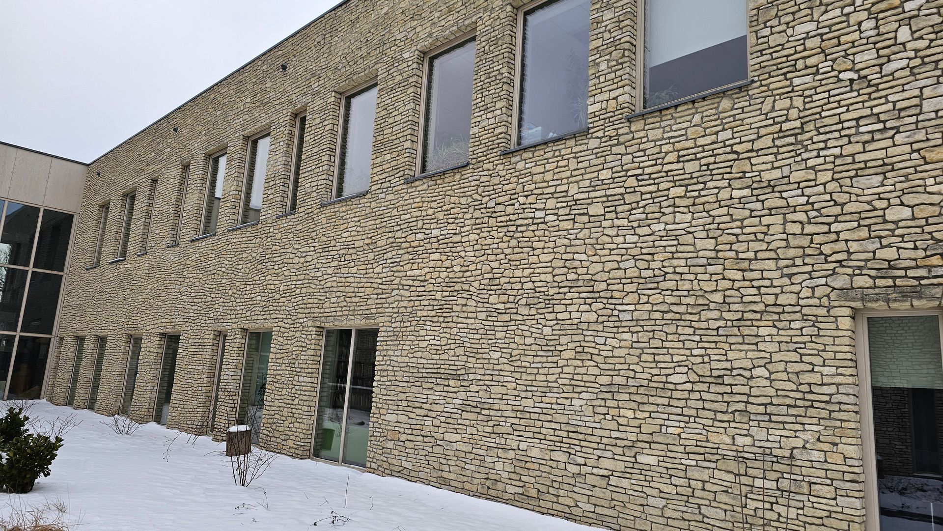 Building with textured stone exterior and multiple windows, snow on the ground.
