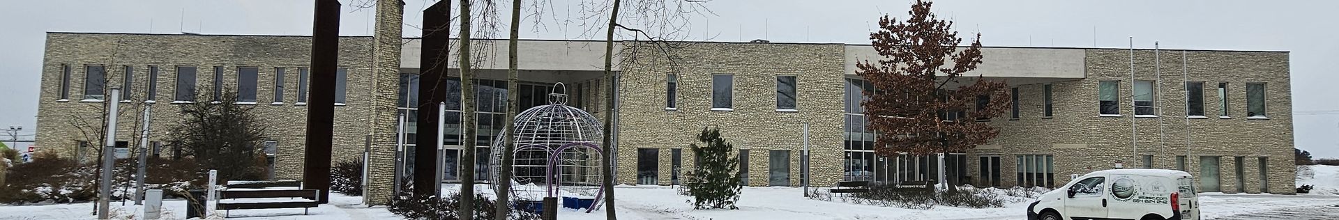 A large stone building with multiple windows sits in a snowy environment with trees. A white van is parked nearby.