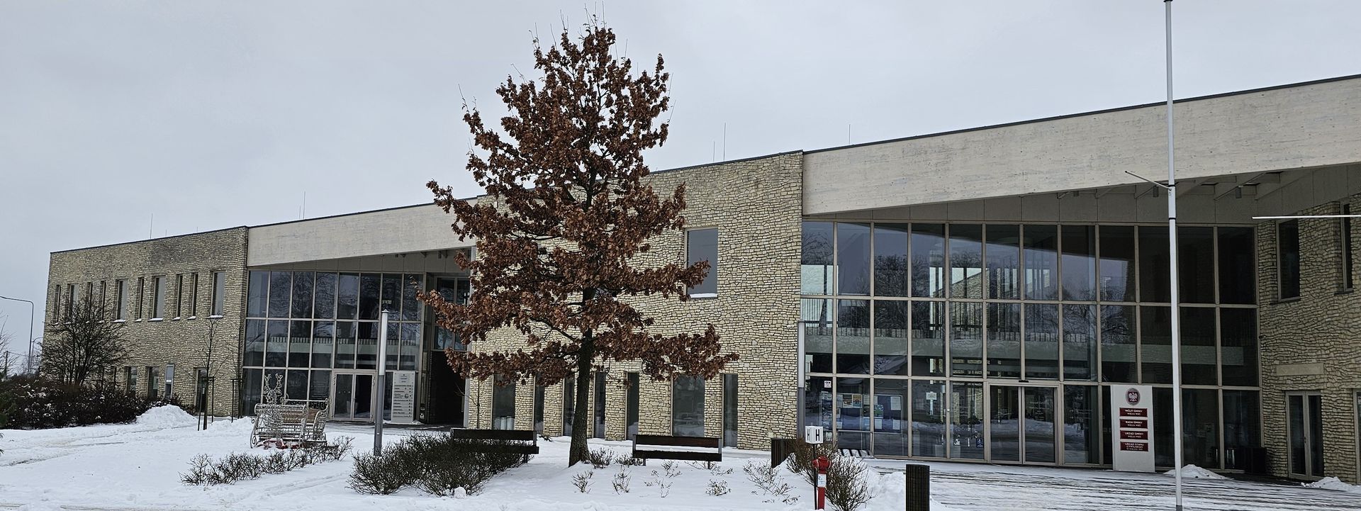 A two-story building with a snow-covered landscape and a leafless tree in front.