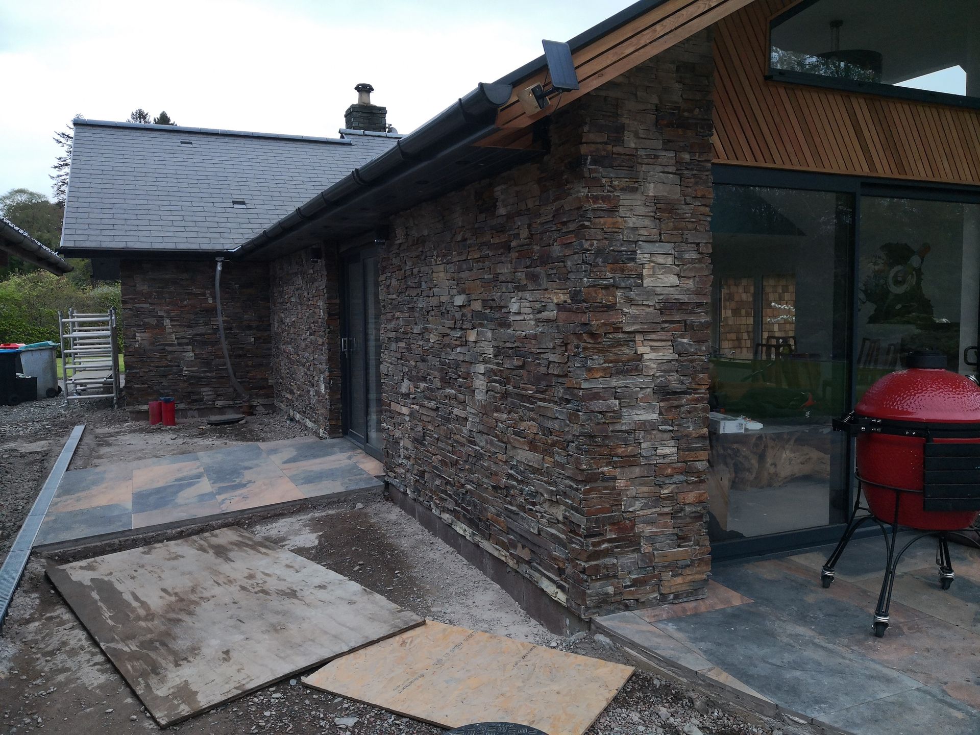 Stone-clad house exterior with dark roof and large windows, a grill on the patio, and construction materials.