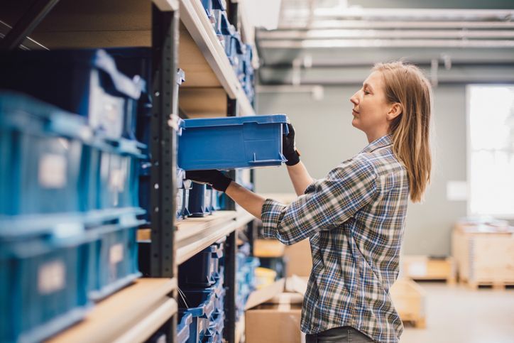 A woman is picking up a blue box from a shelf in a warehouse.
