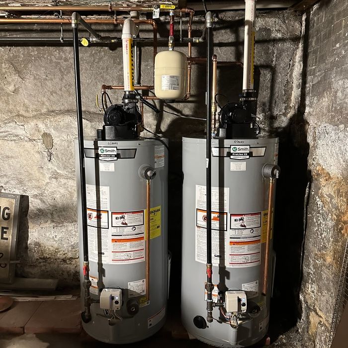 Two gray water heaters with pipes and valves in a basement utility room