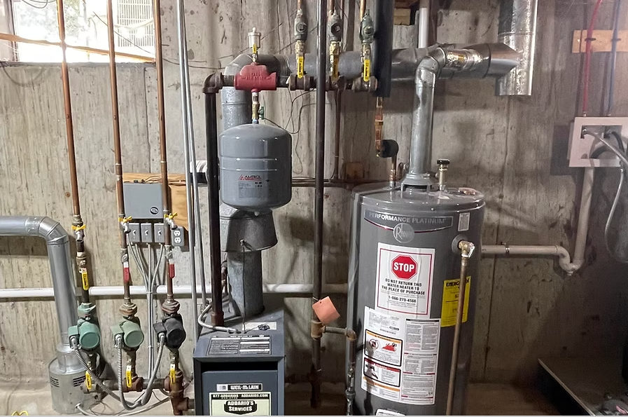 Basement utility area with water heater, pump, pipes, and wall-mounted electrical outlet.