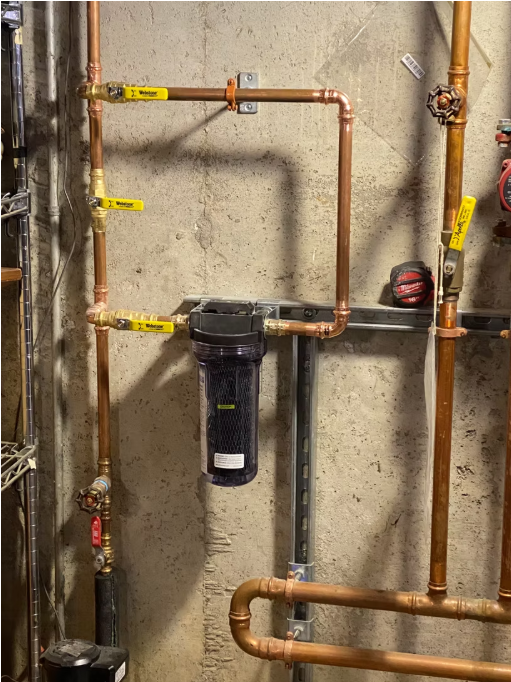 Basement utility wall with copper pipes and a mounted black water filter.