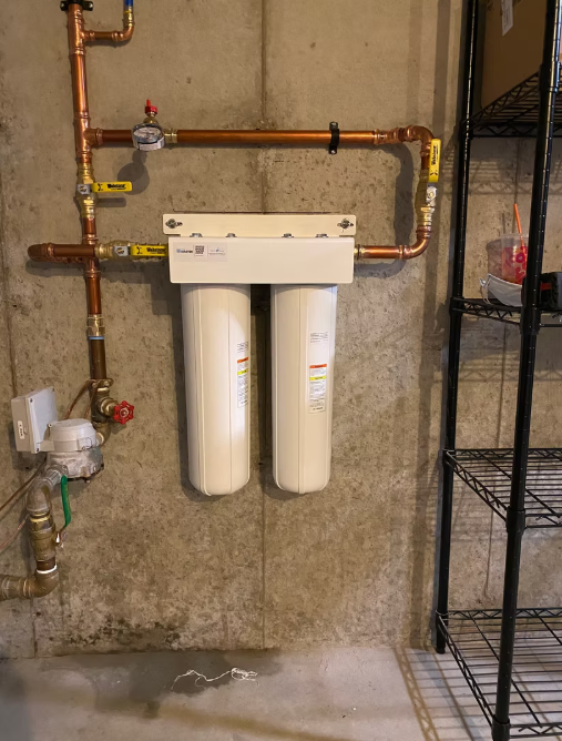 Wall-mounted water filtration system with pipes, two white filter canisters, and shelving in a utility room