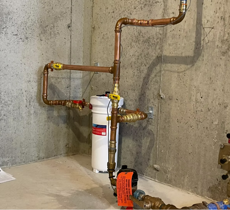 Basement utility pipes with a white water filter mounted on a concrete wall