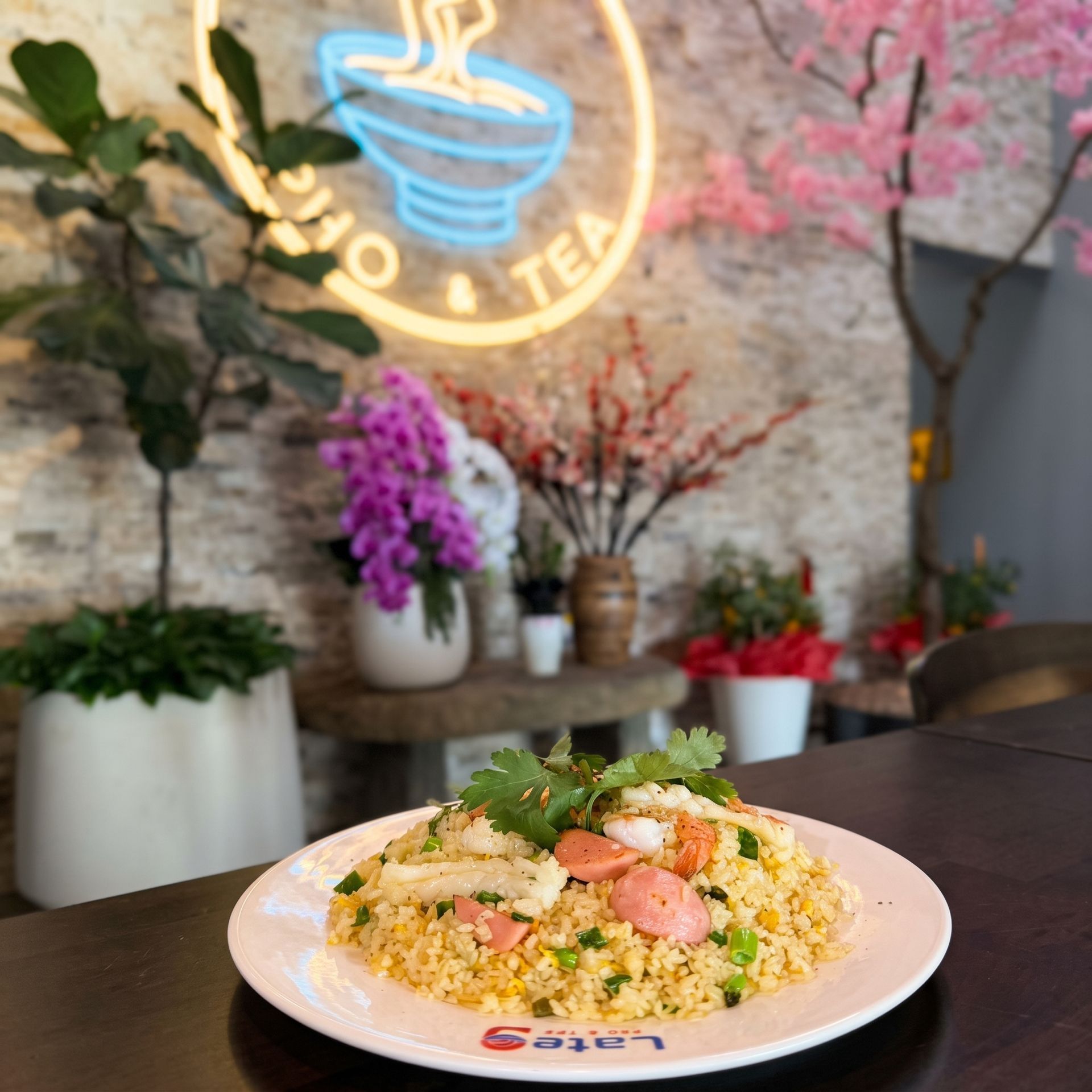Plate of fried rice with shrimp and herbs on a café table, with a neon tea sign and potted plants in back
